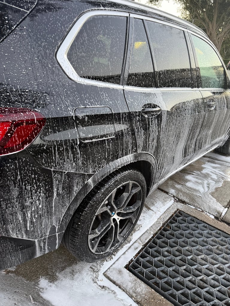 Silver SUV being washed with water spray, covered in soap suds and water droplets during an outdoor car wash