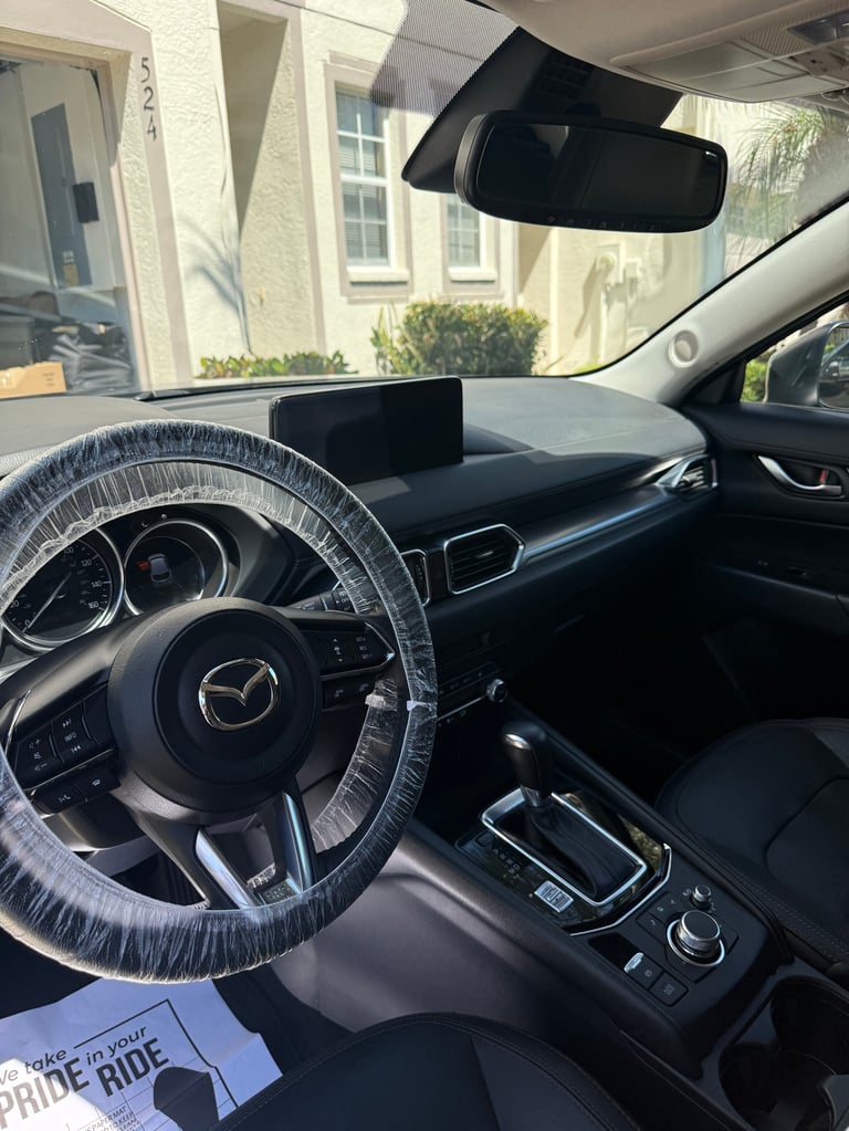 Interior of a Mazda vehicle with clear steering wheel cover, black dashboard, and a building visible through the windshield